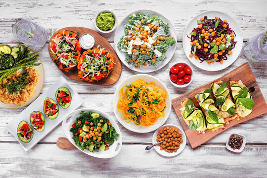 Healthy plant based low carb food table scene. Top down view on a white wood background. Cauliflower flatbread and steak, vegetable noodles, kale salads, stuffed avocados and sweet potatoes.