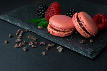 Zwei Pinke Macarons vor dunklem Hintergrund mit Schokosplittern auf Schiefer, mit Himbeere, Minze...