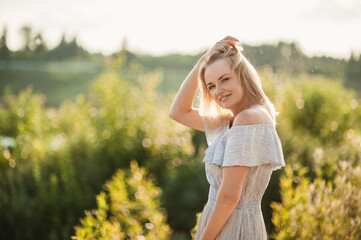 Naklejka na ściany i meble Portrait of a beautiful blonde woman in a summer dress at sunset among the beautiful nature. The girl adjusts her hair and looks into the camera. Space for text.