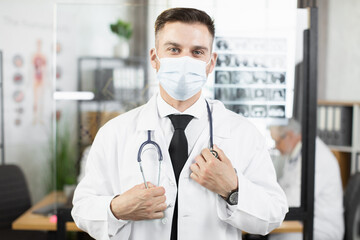 Portrait of confident caucasian doctor in face mask and white lab coat holding stethoscope on neck while standing at workplace. Concept of professional and medicine.