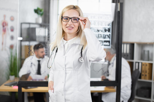 Portrait of beautiful caucasian woman wearing eyeglasses and white lab coat posing at hospital office. Competent female doctor smiling and looking at camera indoors.