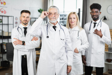 Senior male doctor in eyewer and lab coat standing with his multiracial team at hospital room and looking at camera. Four medical workers posing indoors after meeting.