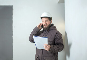 a man in a white construction helmet is standing with documents in the room, and emotionally talking on the phone.