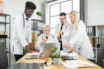 Team of multiracial healthcare experts in white lab coats looking on laptop screen during discussion on meeting. Concept of medicine, conference and technology.