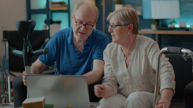 Elder people with walking frame and crutches using laptop in living room. Retired man and woman with chronic disability browsing internet on computer and enjoying technology on free time.