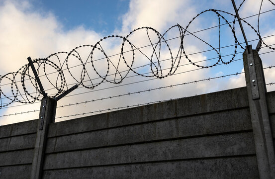 Tall concrete wall topped with razor wire forms a harsh, high-security barrier. The image conveys restriction, safety, and deterrence in an industrial or border environment.