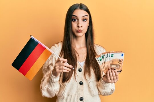 Beautiful brunette young woman holding germany flag and euros banknotes puffing cheeks with funny face. mouth inflated with air, catching air.
