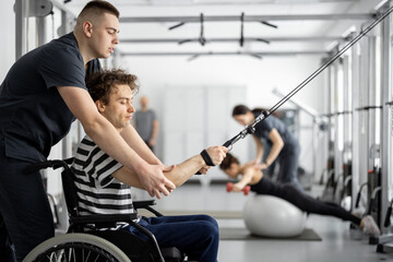 Obraz premium Rehabilitation specialist helps a guy in a wheelchair to do exercise on decompression simulator for recovery from injury. Concept of physical therapy for people with disabilities
