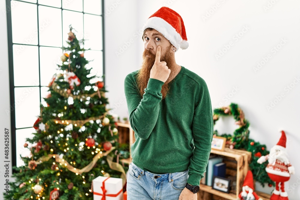 Poster redhead man with long beard wearing christmas hat by christmas tree pointing to the eye watching you