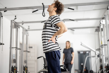 Fototapeta na wymiar Guy feeling back pain while exercising in a rehabilitation center. Concept of physical therapy and kinesiology for muscle recovery and strengthening