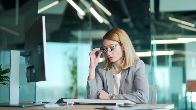 Overworked tired lady holding glasses feeling headache, having eyesight problem after computer work. Stressed mature employee business woman suffering fatigue rubbing dry eyes at workplace in office