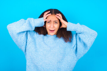 Shocked panic African teenager girl wearing blue sweater over blue background holding hands on head...