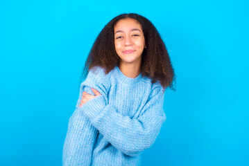 Fototapeta premium People, lifestyle, youth and happiness concept. Shy pretty African teenager girl wearing blue sweater over blue background, feeling happy hugging herself.
