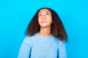 African teenager girl wearing blue sweater over blue background looking up as he sees something strange.