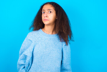 African teenager girl wearing blue sweater over blue background with snobbish expression curving lips and raising eyebrows, looking with doubtful and skeptical expression, suspect and doubt.