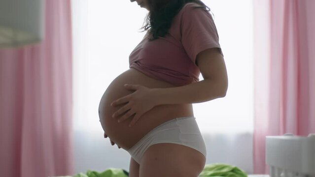 Lonely young pregnant woman is dancing cheerfully in bedroom at home. Happy expecting mother moves gracefully stroking unborn baby in her belly. Motherhood care joy concept