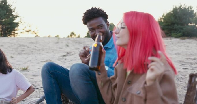 A woman with pink hair and glasses sits on the sand next to boyfriend, singing their favorite song together, holding beer bottles in their hands, having a great time by the fire with friends