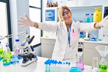 Middle age blonde woman working at scientist laboratory looking at the camera smiling with open arms for hug. cheerful expression embracing happiness.