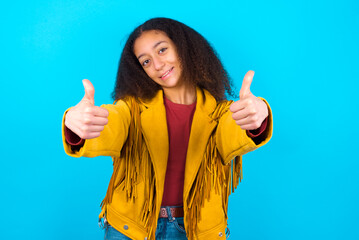 African teenager girl wearing yellow jacket over blue background making positive gesture with thumbs up smiling and happy for success. Looking at the camera, winner gesture.