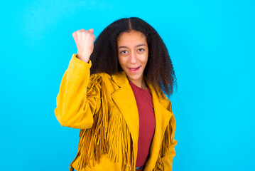 Overjoyed African teenager girl wearing yellow jacket over blue background glad to receive good news, clenching fist and making winning gesture.