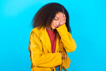 African teenager girl wearing yellow jacket over blue background making facepalm gesture while...