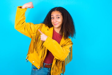 Smiling African teenager girl wearing yellow jacket over blue background raises hand to show muscles, feels confident in victory, strong and independent.
