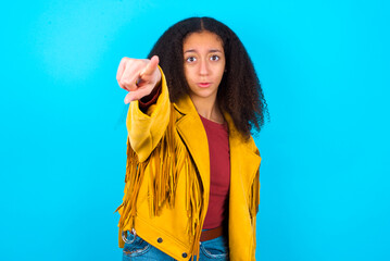 Cheerful African teenager girl wearing yellow jacket over blue background indicates happily at you, chooses to compete, has positive expression, makes choice.