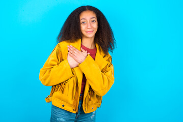 Honest African teenager girl wearing yellow jacket over blue background keeps hands on chest,...