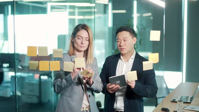 Startup and implementation of a small business project. Asian Businessman discussing with businesswoman sticking adhesive notes on glass wall in office, discussing ideas on visualization board