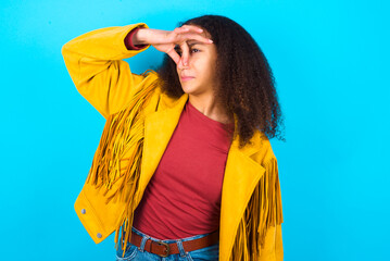 Displeased African teenager girl wearing yellow jacket over blue background plugs nose as smells...