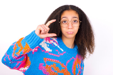 African teenager girl wearing colorful sweater over white background purses lip and gestures with hand, shows something very little.