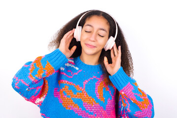 Pleased African teenager girl wearing colorful sweater over white background enjoys listening pleasant melody keeps hands on stereo headphones closes eyes. Spending free time with music