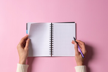 Female hand writing in blank open spiral notebook with pen on pastel pink background. Top view,...