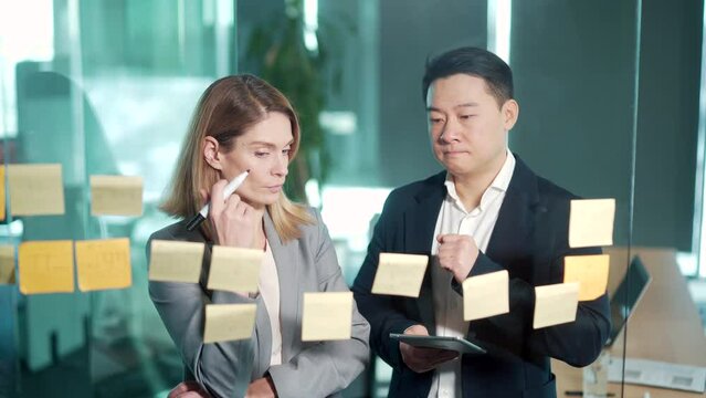 Startup and implementation of a small business project. Asian Businessman discussing with businesswoman sticking adhesive notes on glass wall in office, discussing ideas on visualization board