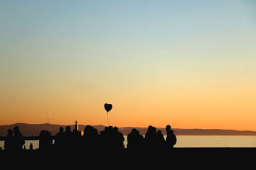 Fototapeta na wymiar Silhouette of unrecognizable group of people and heart shaped balloon, enjoying sunset. Selective focus.