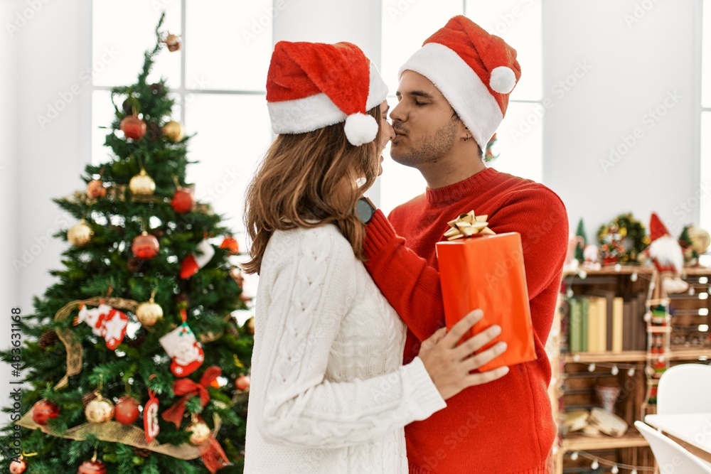 Wall mural young hispanic couple wearing christmas hat holding gift at home.