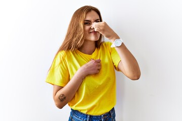 Young brunette woman standing over isolated background smelling something stinky and disgusting, intolerable smell, holding breath with fingers on nose. bad smell - obrazy, fototapety, plakaty