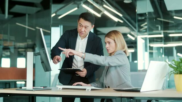 two business people consulting on a project looking at computer monitor screen sitting in the office. Asian male employee Coworker explains colleagues ideas and strategies accountant, financial report