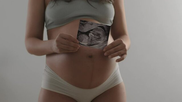 Pregnant woman shows an ultrasound photo of her unborn child to camera. Unrecognizable expecting mom wearing underwear holds a picture of a baby inside her belly