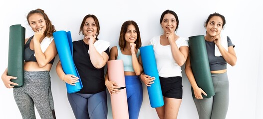 Group of women holding yoga mat standing over isolated background with hand on chin thinking about question, pensive expression. smiling and thoughtful face. doubt concept.