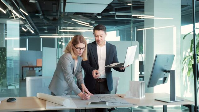 A team of multiracial business architects and designers discuss creating a project draft drawing by looking at laptop monitor screen a computer desk. Asian male and female developers together teamwork