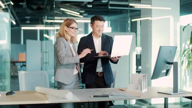A team of multiracial business architects and designers discuss creating a project draft drawing by looking at laptop monitor screen a computer desk. Asian male and female developers together teamwork