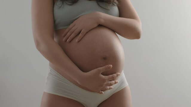Young pregnant woman wearing underwear strokes her belly with care and tenderness. Close-up of expectant mother's stomach on white background