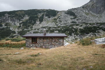 Fototapeta na wymiar Scenic mountain hut made of stone with flowers on the window and a rocky hill in the background