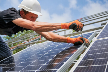 Close up of male worker mounting photovoltaic solar panel system outdoors. Man installer placing solar module on metal rails, wearing construction helmet and work gloves, tightening with hex key.