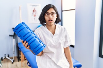 Young hispanic physiotherapist woman holding foam roller at the clinic scared and amazed with open mouth for surprise, disbelief face