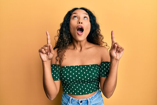 Young latin woman wearing casual clothes amazed and surprised looking up and pointing with fingers and raised arms.