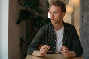 Portrait of young pensive man holding eyeglasses planning project looking at window sitting in cafe 