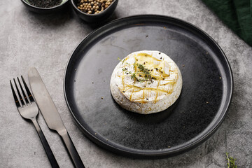 Oven baked camembert cheese with black pepper, sesame seeds on black plate, grey concrete surface. Homemade grilled brie with thyme, dark table setting
