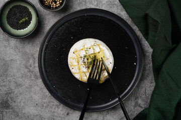 Oven baked camembert cheese with black pepper, sesame seeds on black plate, grey concrete surface. Homemade grilled brie with thyme, dark table setting
